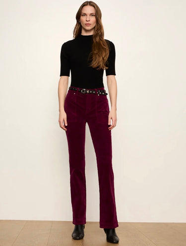 A woman stands against a plain off-white wall wearing a black short-sleeve mock-neck top and deep burgundy corduroy pants with a black studded belt, paired with black ankle boots.