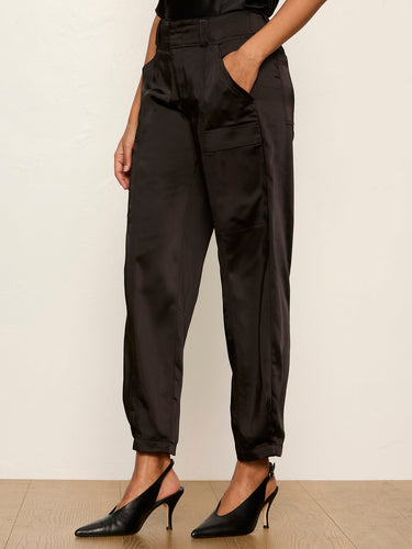 A model wearing black high-waisted satin trousers with side pockets and a cropped ankle length, styled with black slingback heels against a neutral cream wall and light wood floor.