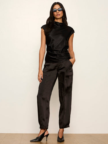 A woman modeling a black satin two-piece set consisting of a short-sleeve mock-neck top and matching wide-leg trousers, paired with black pointed slingback heels against a plain off-white wall.