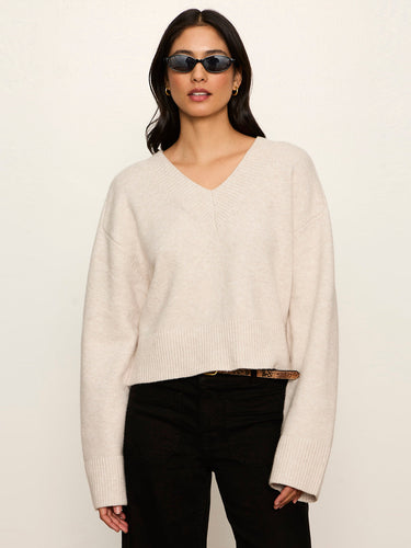 A woman modeling a cream-colored knit V-neck sweater with black pants and sunglasses, standing against a plain off-white background.