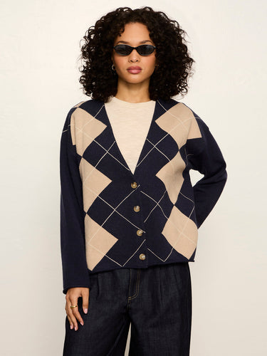 A model with curly black hair and sunglasses wearing a navy blue cardigan with a beige argyle pattern, styled with a cream top and dark jeans against a plain light background.