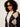 A woman with curly black hair wearing a navy and beige argyle cardigan over a cream top, styled with black sunglasses against a plain light background.