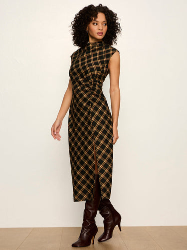 A woman modeling a sleeveless high-neck midi dress with a black and tan plaid pattern, paired with dark brown heeled boots, against a plain off-white studio background.