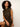 A woman with curly black hair wearing a sleeveless black dress with a tan plaid pattern, sitting against a plain off-white background.