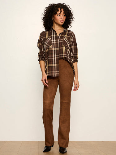 A model stands against a plain off-white wall wearing a brown and beige plaid button-up shirt with gold buttons and brown suede-like straight-leg pants.