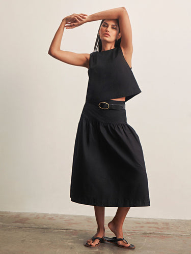 Woman wearing a black sleeveless top and black skirt with a belt against a plain background