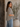 A woman wearing a black and white patterned short-sleeve blouse and light blue jeans, standing in a rustic indoor setting next to a large wooden door and a potted plant.