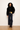 A model stands against a plain off-white wall wearing a black velvet jacket over a black top, dark wide-leg jeans, and brown leather boots, with a yellow plaid shirt tucked into the jeans.