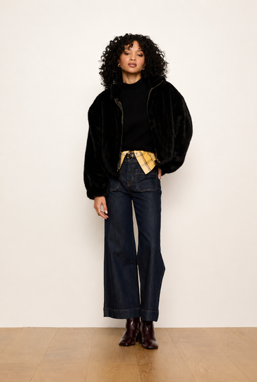 A model stands against a plain off-white wall wearing a black velvet jacket over a black top, dark wide-leg jeans, and brown leather boots, with a yellow plaid shirt tucked into the jeans.
