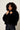 A model with curly dark hair wearing a black cropped faux fur jacket over a black top and dark blue jeans, posing against a light gray studio background.