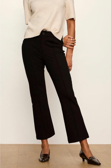 A model wearing black cropped flare pants, paired with a cream short-sleeve top and black pointed-toe heels, standing against a plain off-white wall.