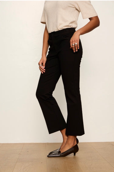 A model wearing black cropped flare pants with a beige short-sleeve top and black pointed-toe flats, standing against a plain off-white wall.