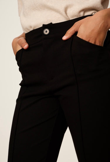 Close-up of a person wearing black trousers with hands in the pockets, showing the waistband with a silver button and part of a light beige top against a plain light background.