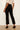 A model shown from the back wearing black cropped ankle-length trousers, a cream short-sleeve top, and black patent leather kitten heels against a plain off-white wall and light wood floor.