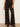 A model wearing high-waisted, wide-leg plaid trousers in a dark brown and black tartan pattern, paired with black leather shoes, standing against a plain off-white wall.