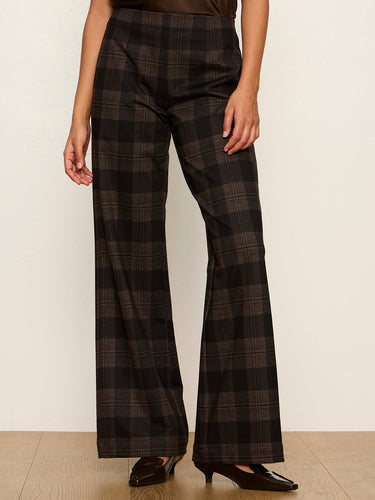 A model wearing high-waisted, wide-leg plaid trousers in a dark brown and black tartan pattern, paired with black leather shoes, standing against a plain off-white wall.