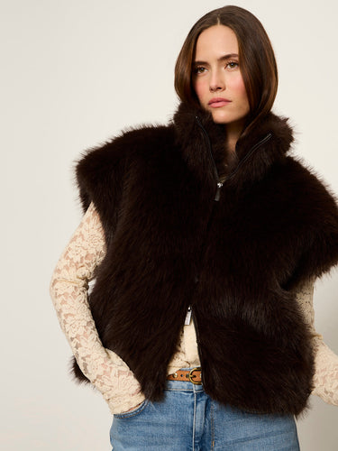 A woman wearing a dark brown fur vest over a cream lace long-sleeve top and blue jeans, photographed against a plain light background.