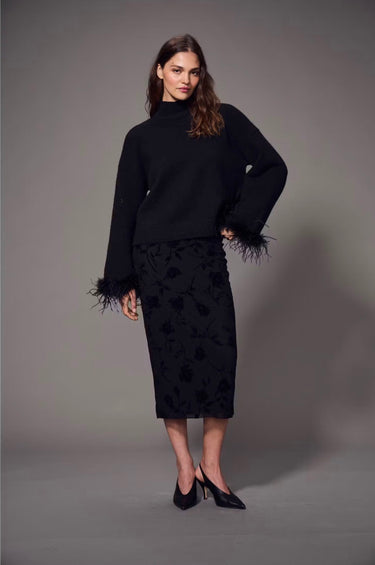 a woman wearing a black sweater with feather cuffs, a mesh midi skirt with a velvet floral pattern and black heels on a grey studio background