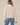 A woman wearing a ribbed beige jacket and blue jeans on a neutral background.