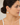 A close-up side profile of a woman wearing a delicate gold hoop earring with a small, semi-circular white stone or marble inlay, paired with a fine gold chain necklace.