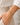 A close-up of a person's wrist wearing a stack of delicate bracelets, including a gold chain with a small gold tassel charm and a silver-toned beaded bracelet, paired with a white blouse and beige trousers.