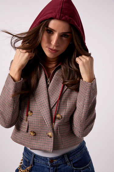 A woman wearing a cropped houndstooth blazer in brown and beige with gold buttons, a burgundy hood, and blue jeans, posing against a light gray studio background.