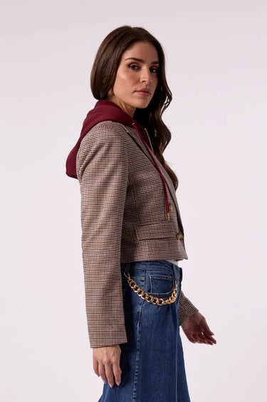 A woman modeling a cropped, long-sleeve plaid jacket with a built-in burgundy hood, paired with blue jeans and a gold chain belt, against a plain light gray background.