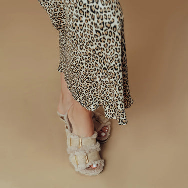 A close-up of a person's foot wearing fuzzy beige double-strap sandals with gold buckle details, paired with a leopard-print skirt, against a solid beige background.