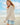Woman wearing a beige knit sweater and denim shorts on a beach with a blue sky.