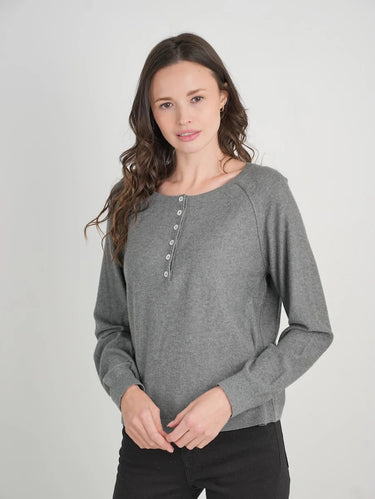 A woman modeling a heather gray long-sleeve henley top with a button placket, paired with black pants, against a light gray studio background.