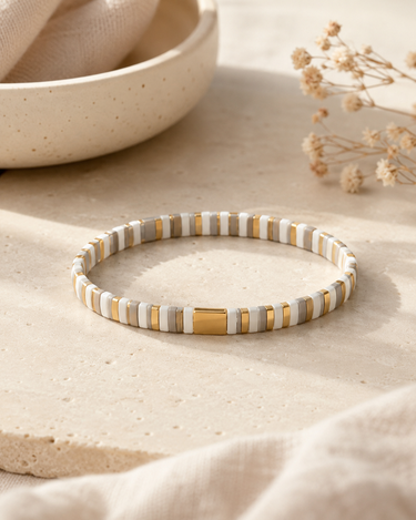 A slim beaded bracelet with alternating white and gold ceramic beads, displayed on a textured beige stone surface with a ceramic bowl and dried flowers in the background.