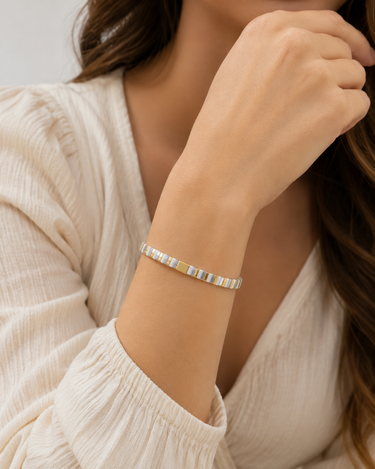 A close-up of a woman wearing a delicate silver bangle bracelet with small gold and white inlay details, paired with a cream-colored textured blouse.