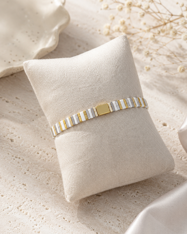 A two-tone silver and gold ceramic bracelet displayed on a cream-colored fabric pillow, set against a textured beige background with dried floral elements.
