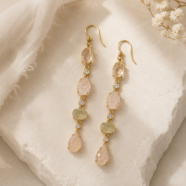A pair of gold drop earrings with peach-pink and light green gemstone beads and small clear stones, displayed on a textured beige stone surface with soft fabric and dried flowers.