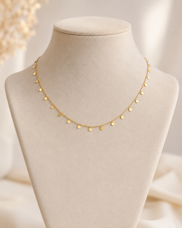 A delicate gold chain necklace with small, evenly spaced flat circular disc charms, displayed on a beige fabric jewelry bust.