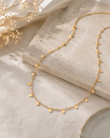 A delicate gold chain necklace with small flat circular disc charms, styled on a textured stone slab and cream fabric with dried flowers.