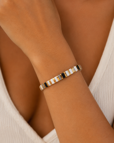 A close-up of a person's wrist wearing a thin beaded bracelet with alternating white, black, and gold metallic beads.