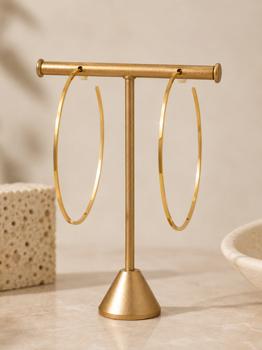 A pair of large gold hoop earrings displayed on a matching gold stand, set against a warm beige background with subtle decorative props.