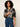 A woman with curly dark hair wearing a long-sleeve plaid button-up shirt in navy, gray, and white, paired with blue jeans, against a plain light background.