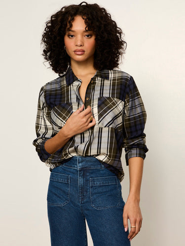 A woman with curly dark hair wearing a long-sleeve plaid button-up shirt in navy, gray, and white, paired with blue jeans, against a plain light background.