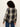 A woman with curly dark hair is shown from the back wearing a long-sleeve plaid button-up shirt in black, white, and blue, paired with blue jeans, against a plain off-white background.