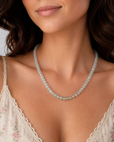 A close-up of a woman wearing a delicate beaded necklace with pale translucent beads and a small cluster of iridescent accent beads at the center, paired with a cream lace-trim camisole.