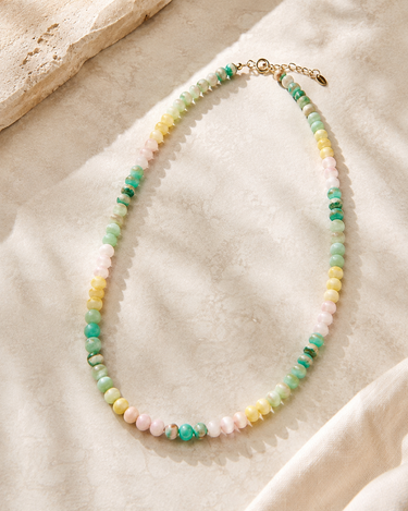 A beaded necklace made of small pastel and green gemstone-like beads with gold-tone clasp, arranged on a soft cream fabric with a stone prop in the corner.