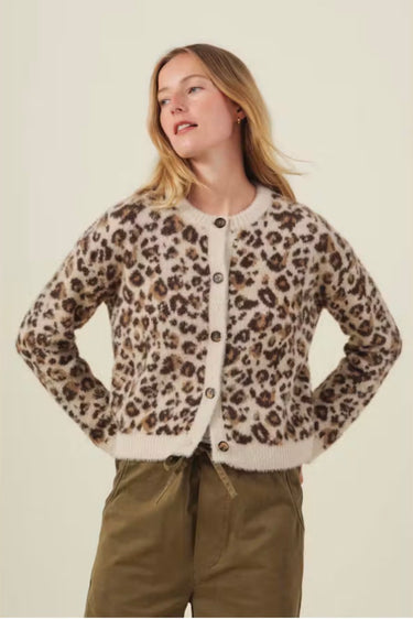 A woman modeling a fuzzy leopard-print cardigan with brown buttons, paired with olive green pants, against a solid light beige background.