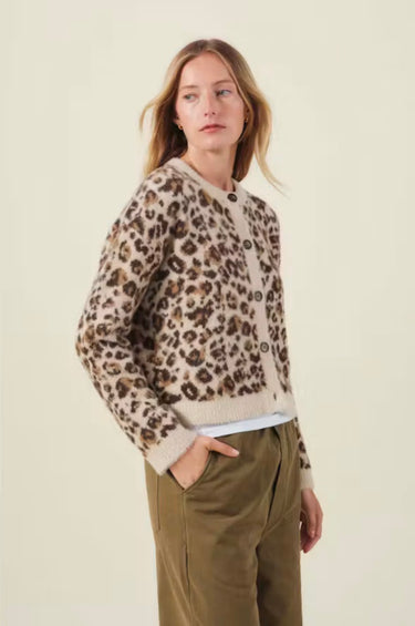 A woman modeling a beige and brown leopard-print knit cardigan with a button-up front, paired with olive green trousers, against a solid light beige background.