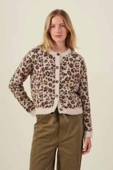 A woman modeling a fuzzy leopard-print cardigan with brown buttons, paired with olive green pants, against a plain cream background.