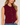 A woman wearing a sleeveless ribbed knit vest in deep burgundy with dark buttons and two chest flap pockets, paired with blue jeans against a light neutral background.