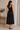 Woman wearing a black dress standing in a room with a wooden bench and plant.