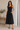 Woman wearing a black dress standing in a minimalistic room with a wooden bench and plant.