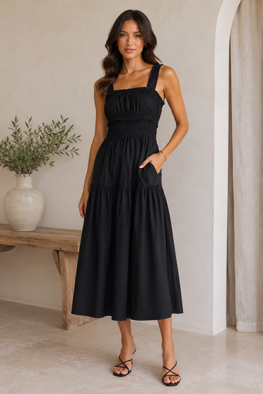 Woman wearing a black dress standing in a minimalistic room with a wooden bench and plant.